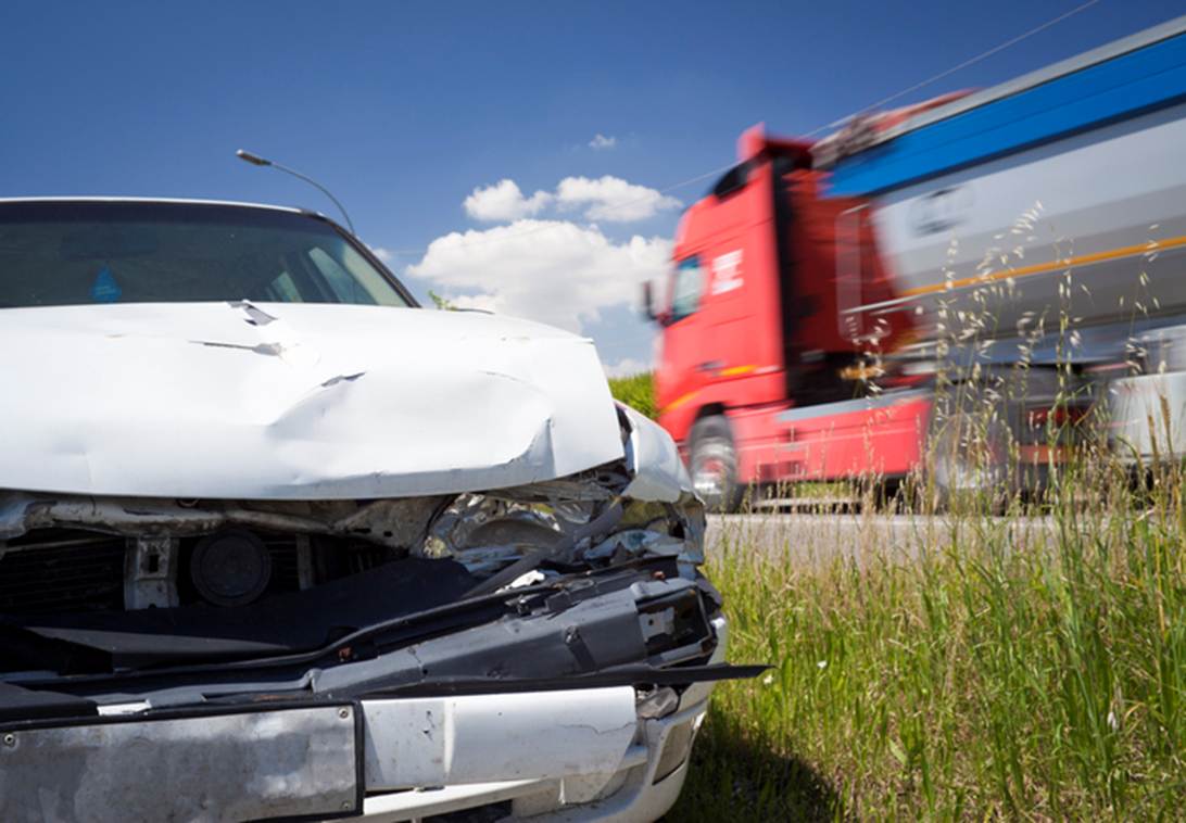This image shows a car crash. The front of a white car is heavily damaged with a red tractor trailer blurred in the background.